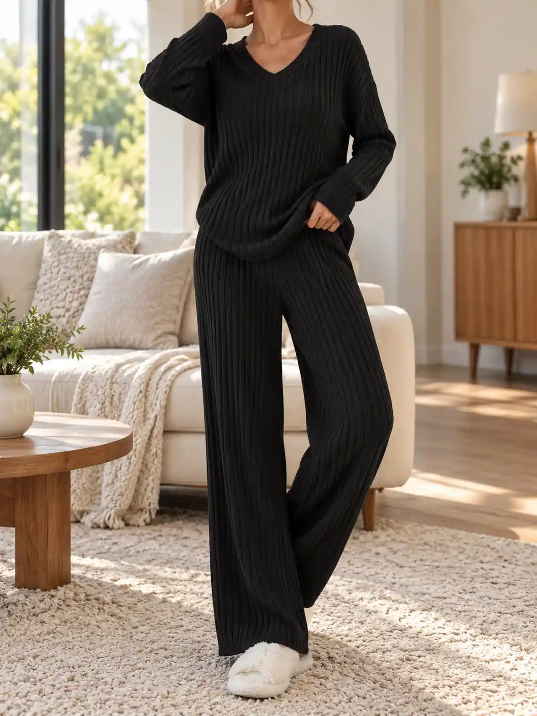 A woman wearing a black ribbed knit lounge set consisting of a long-sleeve V-neck top and matching wide-leg pants, standing in a bright living room with a sofa, rug, and wooden furniture.