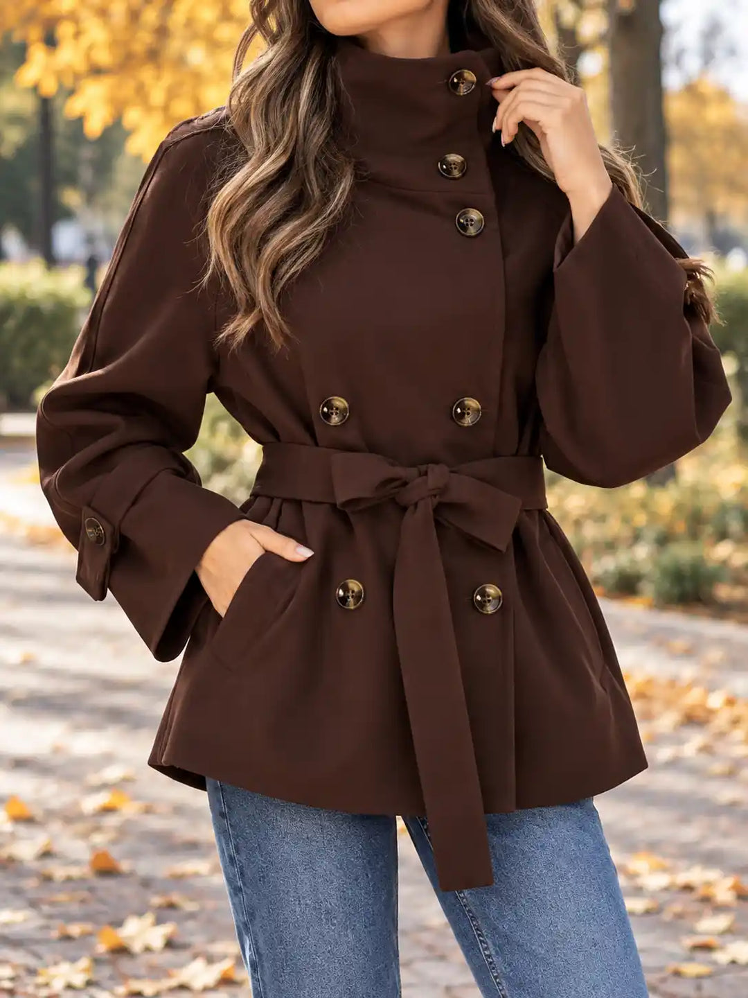 A woman wearing a dark brown double-breasted belted coat with gold-tone buttons, paired with blue jeans, standing outdoors on a path with fallen leaves and trees in the background.