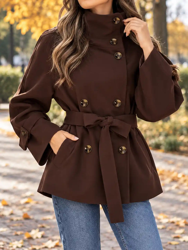 A woman wearing a dark brown double-breasted belted coat with gold-tone buttons, paired with blue jeans, standing outdoors on a path with fallen leaves and trees in the background.