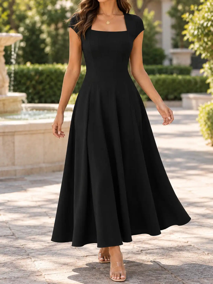 A woman wearing a black midi dress with a square neckline, cap sleeves, and a flared skirt, standing outdoors on a stone walkway with a garden and fountain in the background.