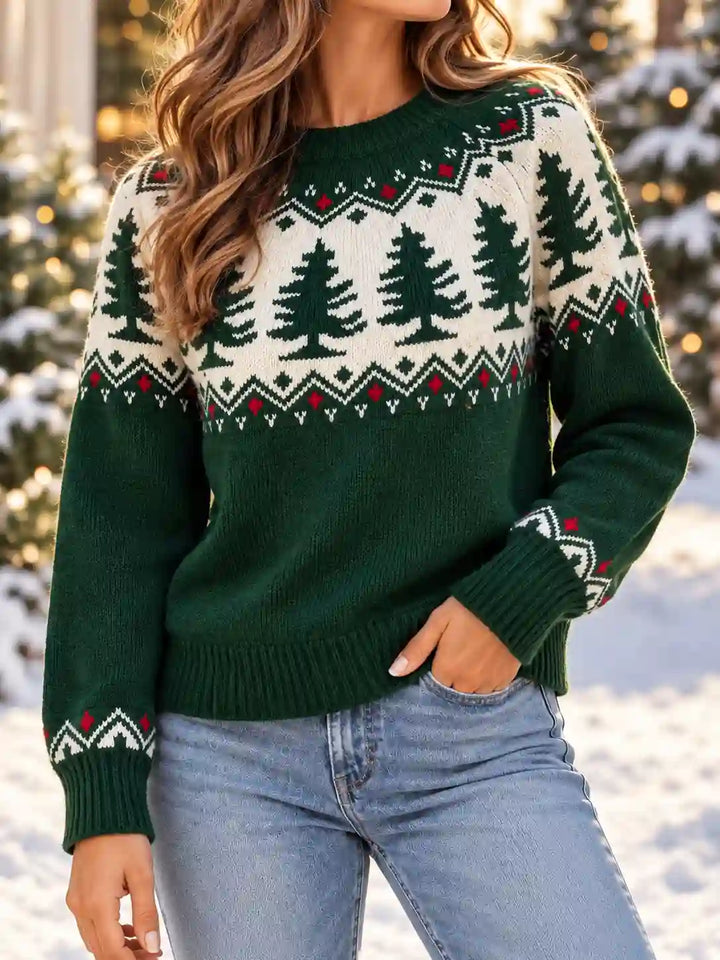 A woman wearing a dark green and cream knit sweater with a Christmas tree pattern, paired with light blue jeans, set against a snowy outdoor background with blurred lights.