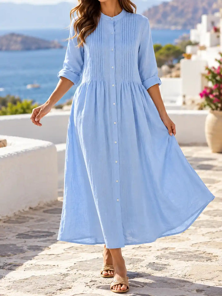 A woman wearing a light blue midi dress with pleated details on the bodice and a button-down front, walking outdoors on a stone path with a scenic blue sea and white buildings in the background.