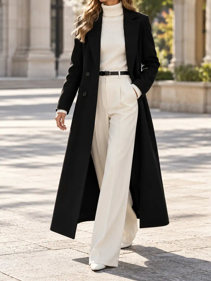 A woman wearing a long black coat over a cream ribbed turtleneck and cream wide-leg trousers, walking outdoors on a sunlit stone walkway.