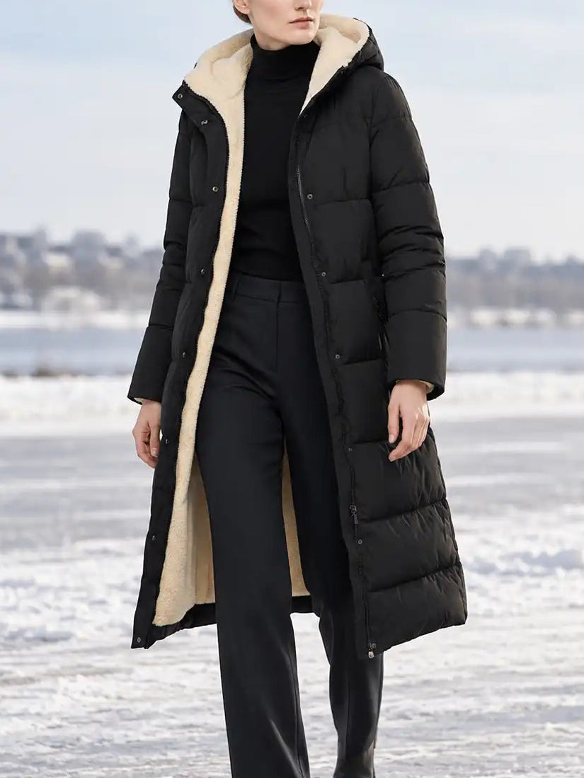 A woman stands outdoors on a snowy, icy surface wearing a long black puffer coat with a beige sherpa-style lining, black trousers, and black boots.