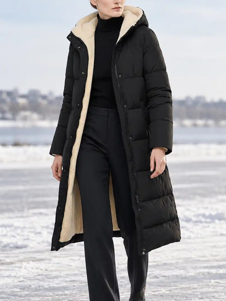 A woman stands outdoors on a snowy, icy surface wearing a long black puffer coat with a beige sherpa-style lining, black trousers, and black boots.