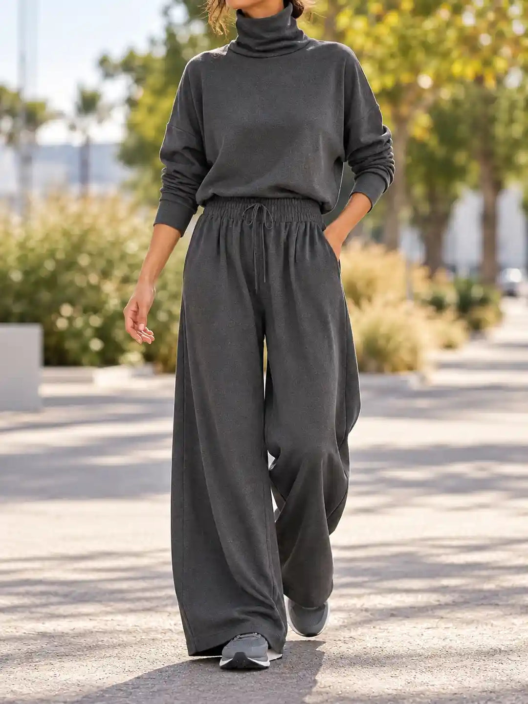 A woman wearing a dark charcoal gray turtleneck sweater and matching wide-leg lounge pants, walking outdoors on a paved path with trees and shrubs in the background.