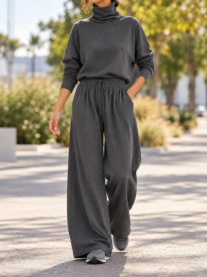 A woman wearing a dark charcoal gray turtleneck sweater and matching wide-leg lounge pants, walking outdoors on a paved path with trees and shrubs in the background.