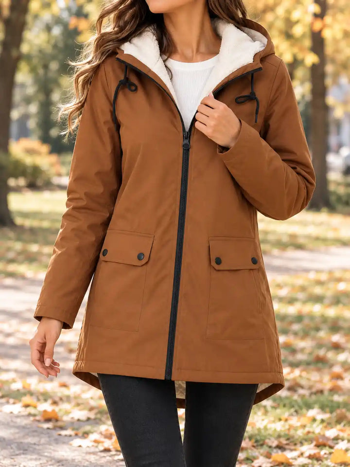 A woman wearing a brown hooded parka jacket with a white sherpa-lined hood and black zipper, standing in an outdoor park setting with autumn trees and fallen leaves.