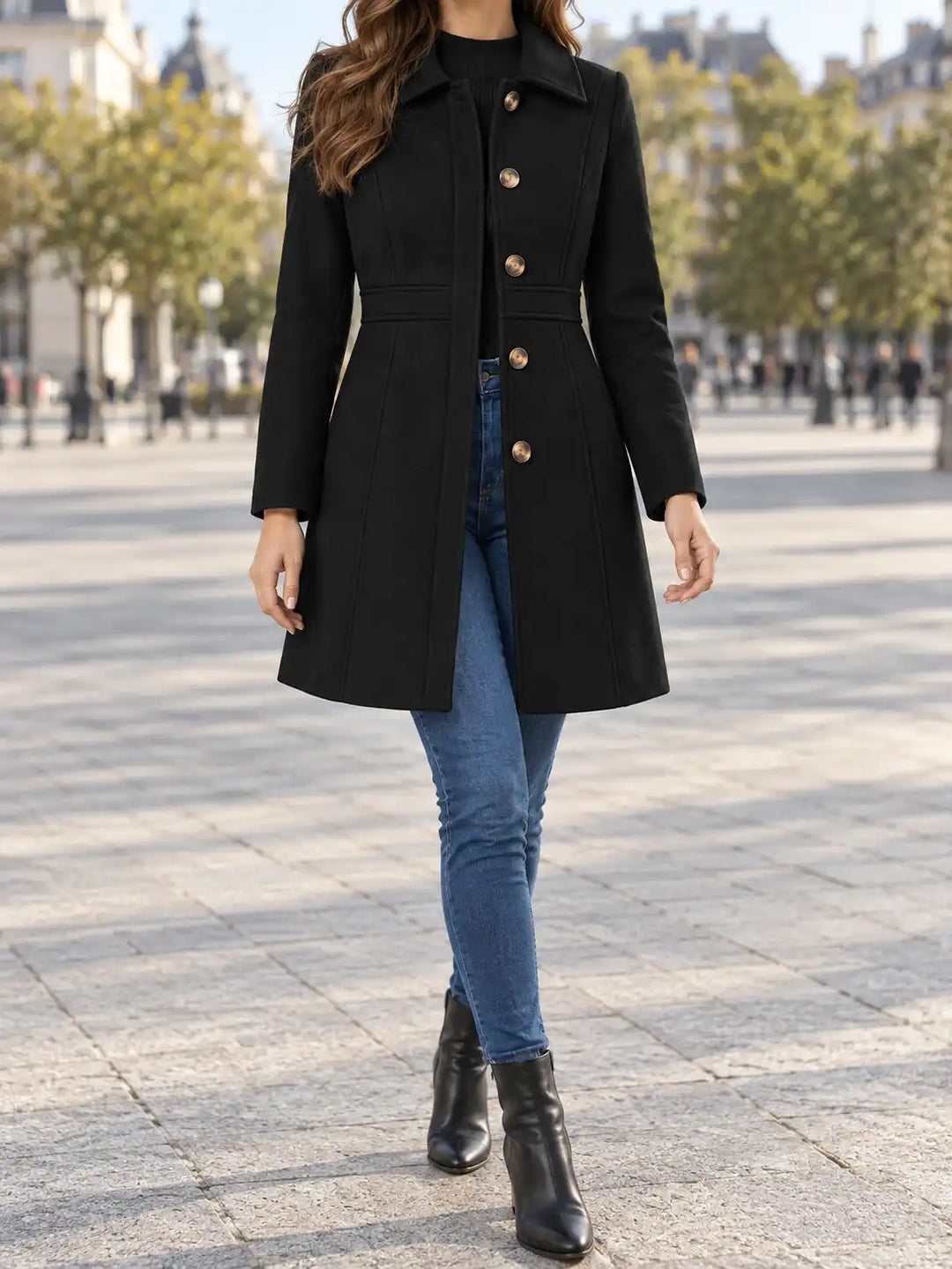 A woman wearing a black mid-length coat with gold buttons, blue jeans, and black ankle boots, walking in a paved outdoor plaza.